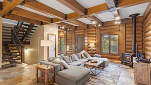 L’intérieur d’une cabane en rondins, avec un escalier en bois, un canapé d’angle, une cheminée et une grande fenêtre.