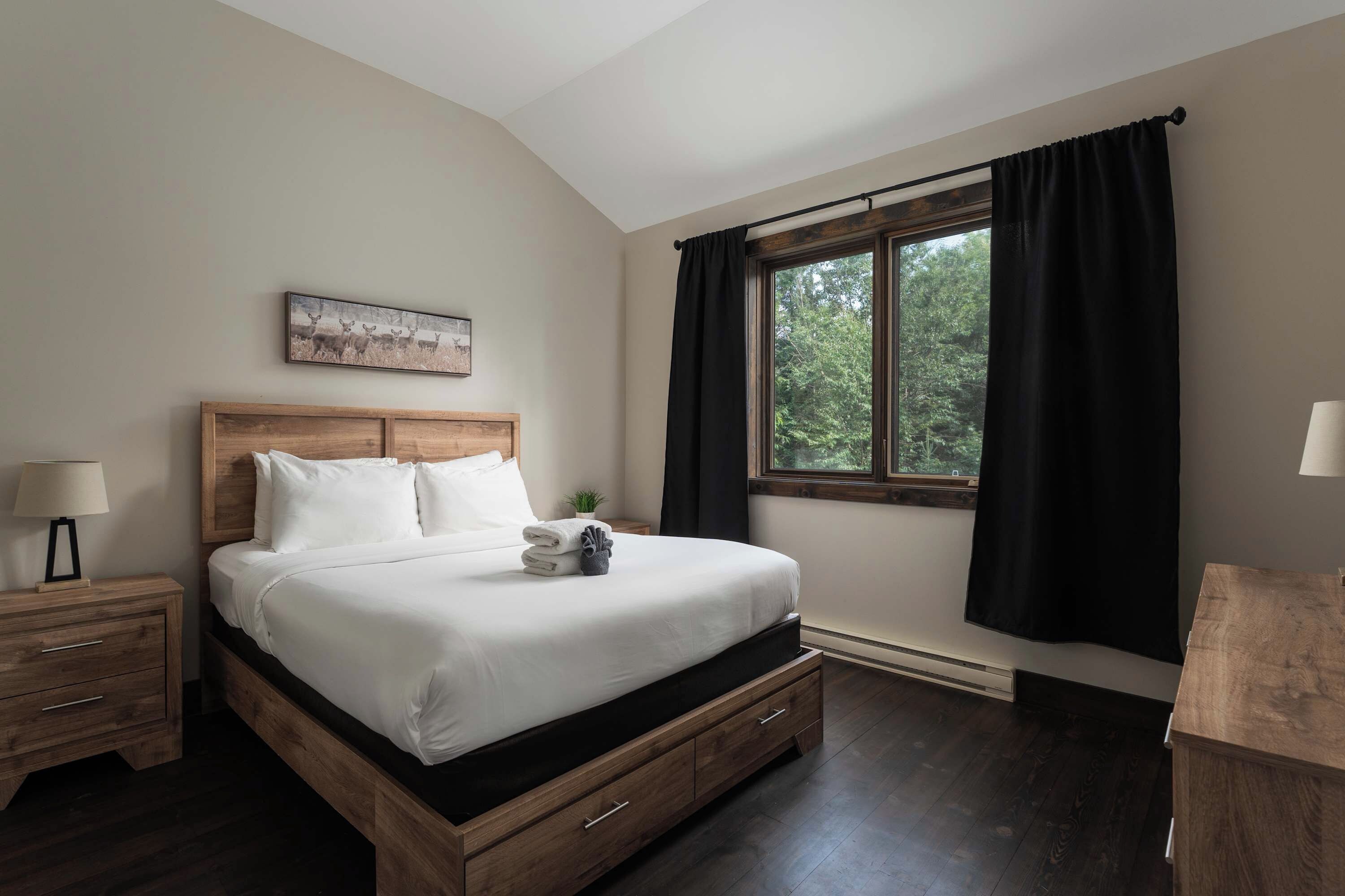 Un dormitorio con una cama de madera, una mesita de noche, una lámpara en la mesita, una ventana con cortinas y un cuadro en la pared.
