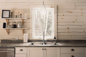 A kitchen with a double sink, wooden shelves, and a window with a view of trees. - Spa, Climbing Wall & Arcade ~ Biking & Golf! (Saint-Faustin-Lac-Carré)