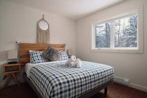 A bedroom with a wooden headboard, a checkered bedspread, a macrame wall hanging, and a window with a view of trees. - Spa, Climbing Wall & Arcade ~ Biking & Golf! (Saint-Faustin-Lac-Carré)