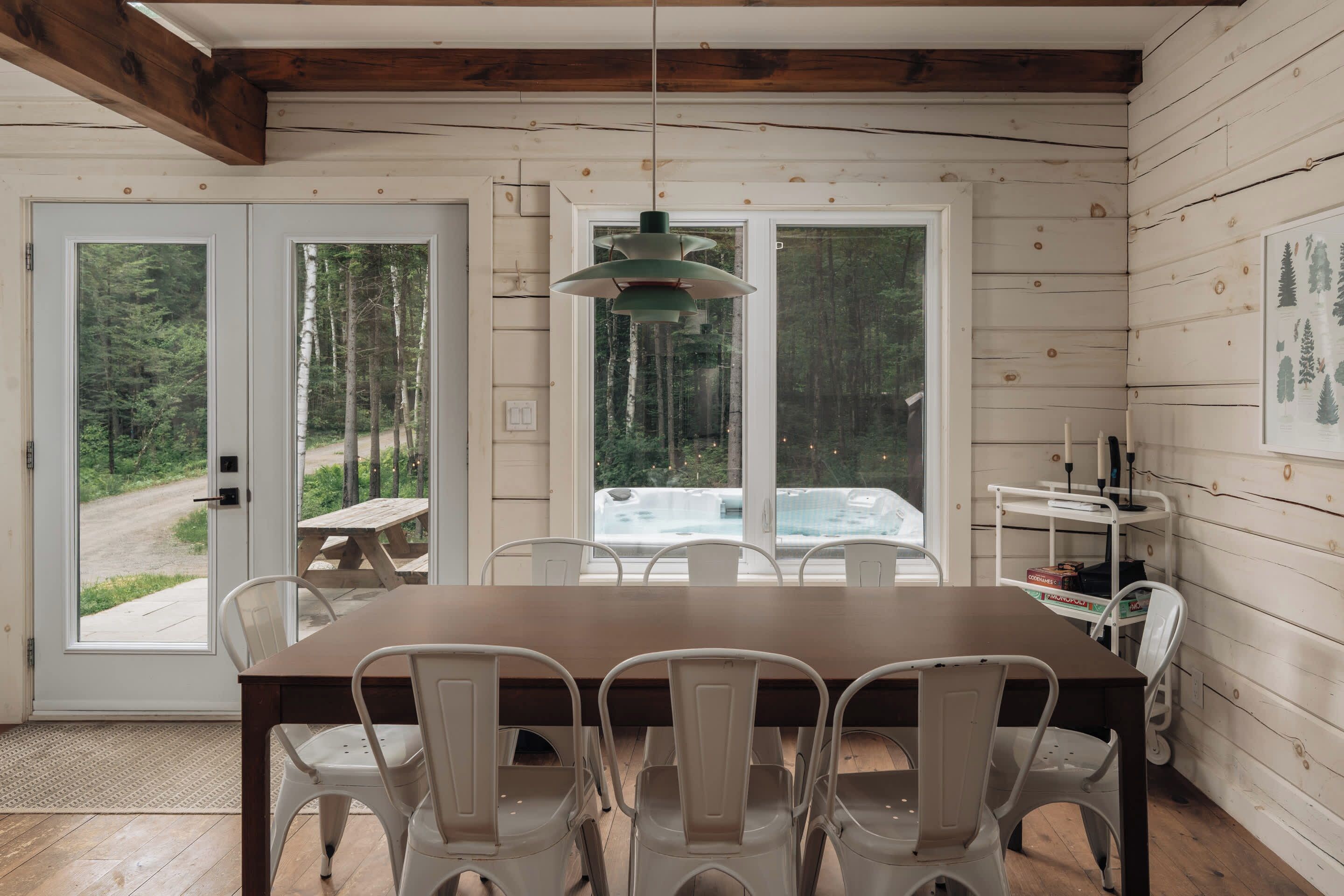 Interno di una baita in legno con un tavolo da pranzo, sedie, un lampadario e vista su una foresta attraverso ampie finestre.