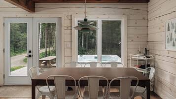 Interior de una cabaña de madera con una mesa de comedor, sillas, una lámpara colgante y vistas a un bosque a través de amplios ventanales.