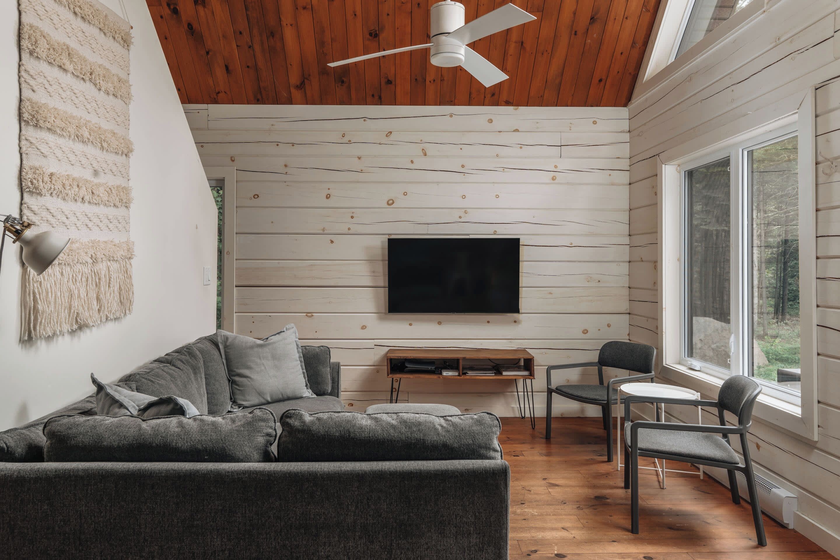 A modern living room with a wooden ceiling, a flat-screen TV mounted on the wall, a gray sofa, and a small table with chairs.