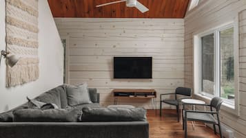 A modern living room with a wooden ceiling, a flat-screen TV mounted on the wall, a gray sofa, and a small table with chairs.