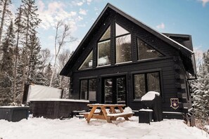 A black wooden cabin with large windows, a picnic table, and a snow-covered landscape. - Spa, Climbing Wall & Arcade ~ Biking & Golf! (Saint-Faustin-Lac-Carré)
