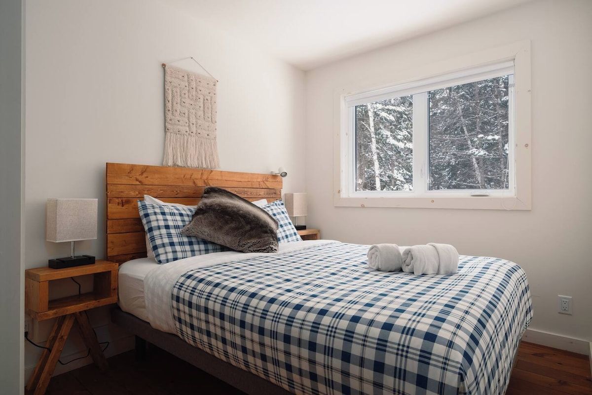 Ein Schlafzimmer mit einem hölzernen Kopfende, einer karierten Tagesdecke, einer Nachttischlampe und einem Fenster mit Blick auf schneebedeckte Bäume.
