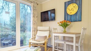 Een kamer met een groot raam, een aan de muur bevestigde televisie, een schilderij, een eettafel met stoelen, en een klein krukje.