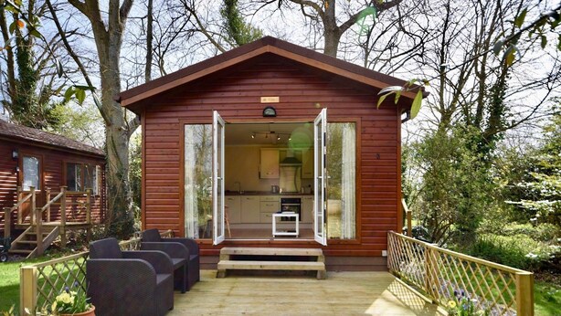 Een houten vakantiehuisje met een openstaande deur naar een keuken, een terras met tuinmeubilair en een omliggende tuin.
