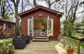 Eine Holzhütte mit offener Tür, die eine Küche zeigt, eine Terrasse mit Gartenmöbeln und einen umlaufenden Garten.