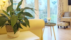 A living room with a yellow sofa, a potted plant, and a small round table with a vase of flowers.