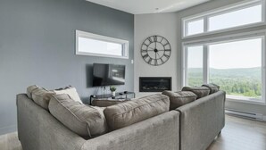 Una sala de estar moderna con un sofá modular, un televisor de pantalla plana, una chimenea y ventanas amplias que ofrecen vistas al exterior.