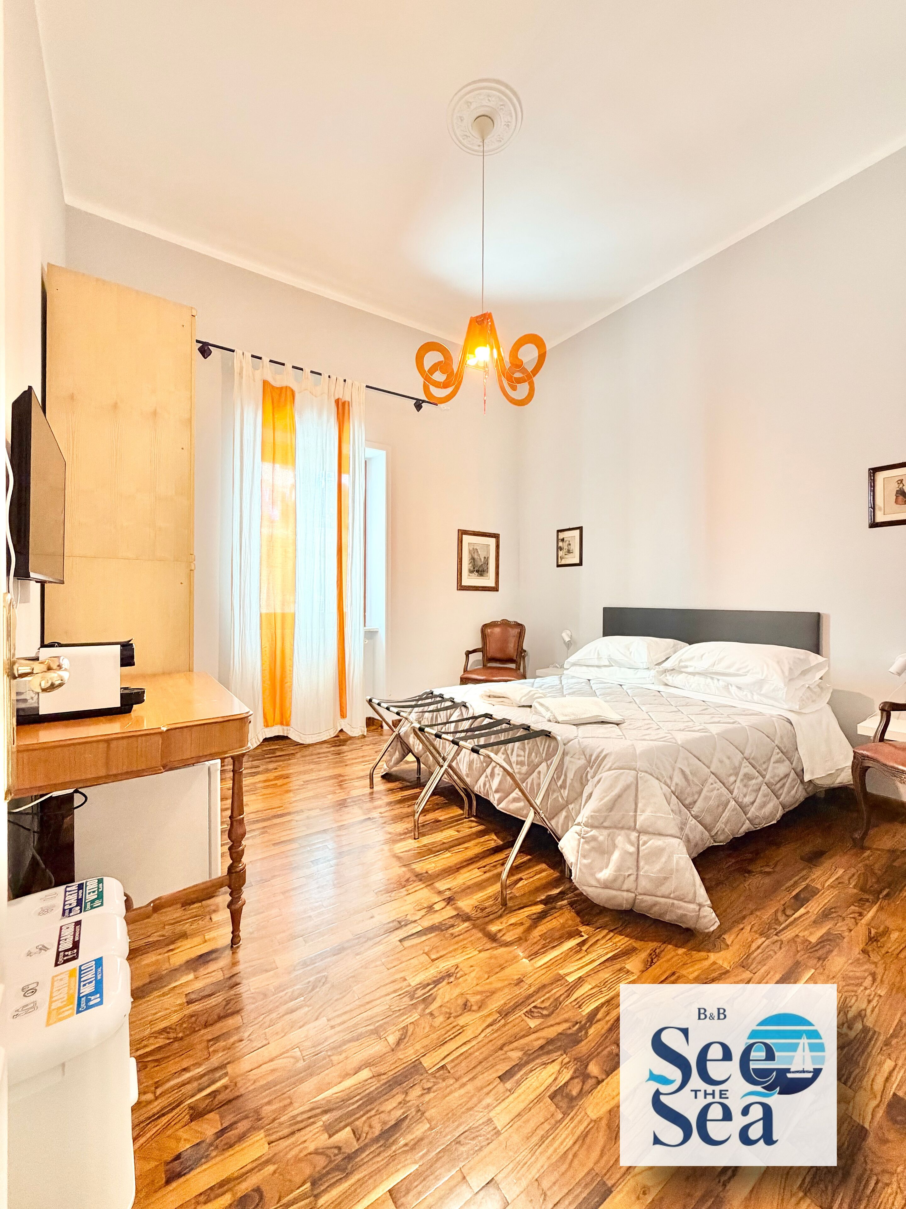 A bedroom with a bed, a desk, a chair, a hanging light fixture, and a window with curtains.