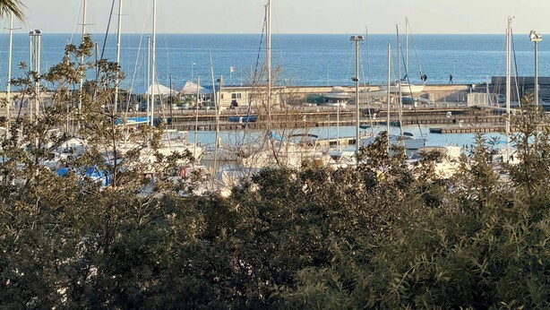 Uma marina com barcos atracados, céu claro e vista distante do mar.