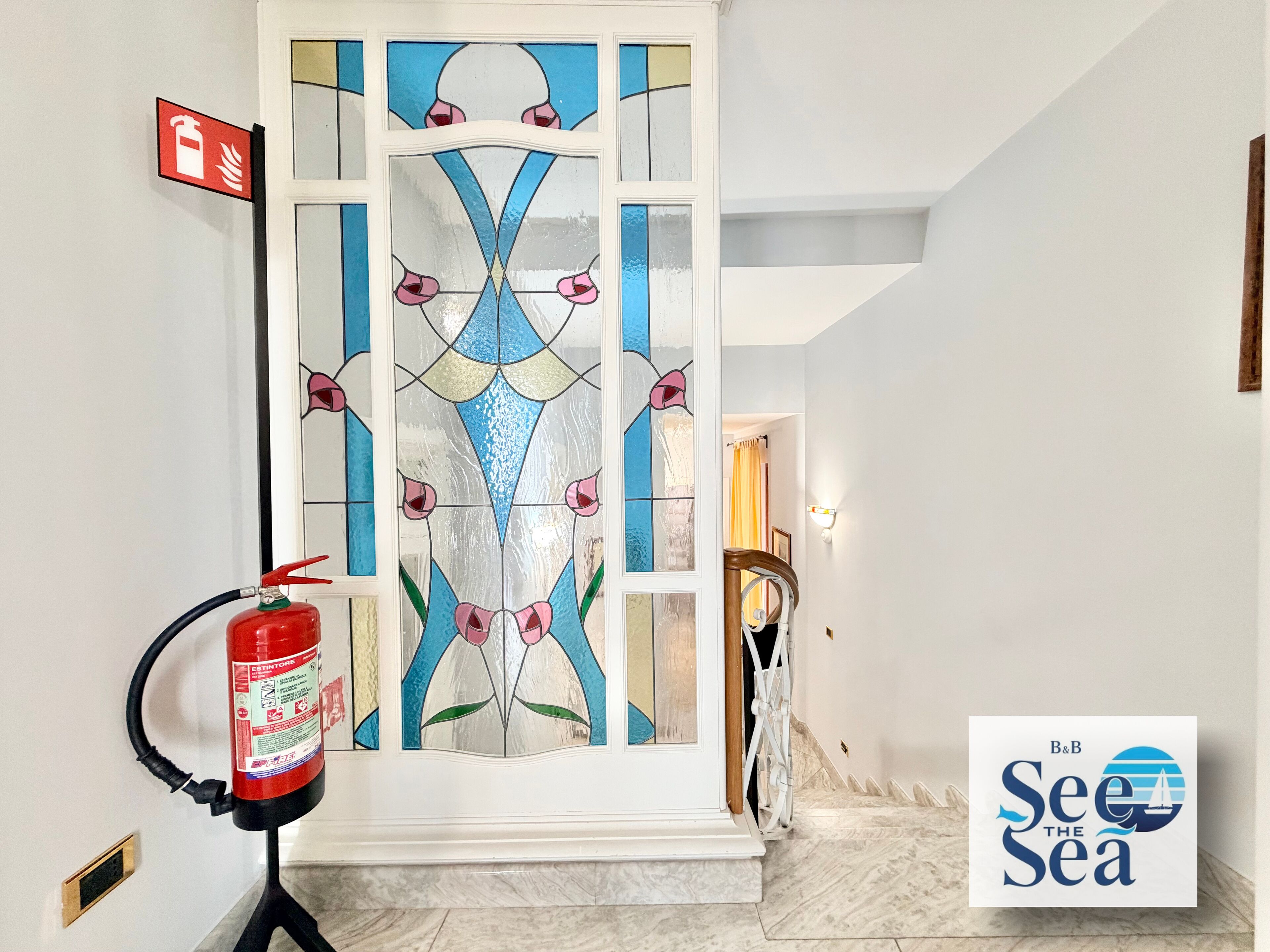 A hallway with a fire extinguisher, a glass door with a floral pattern, and a sign reading 'B&B See the Sea'.