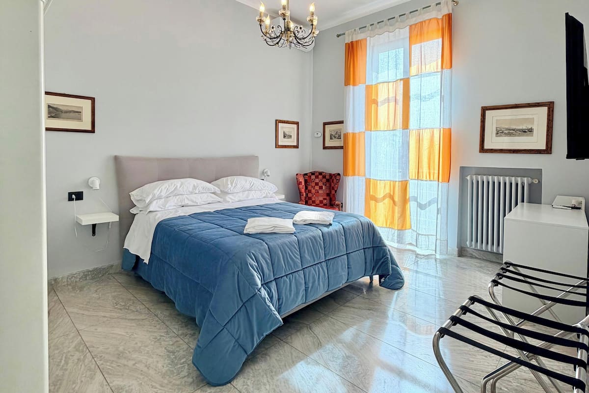 A bedroom with a bed, a chandelier, a window with curtains, a radiator, a chair, and framed pictures on the walls.