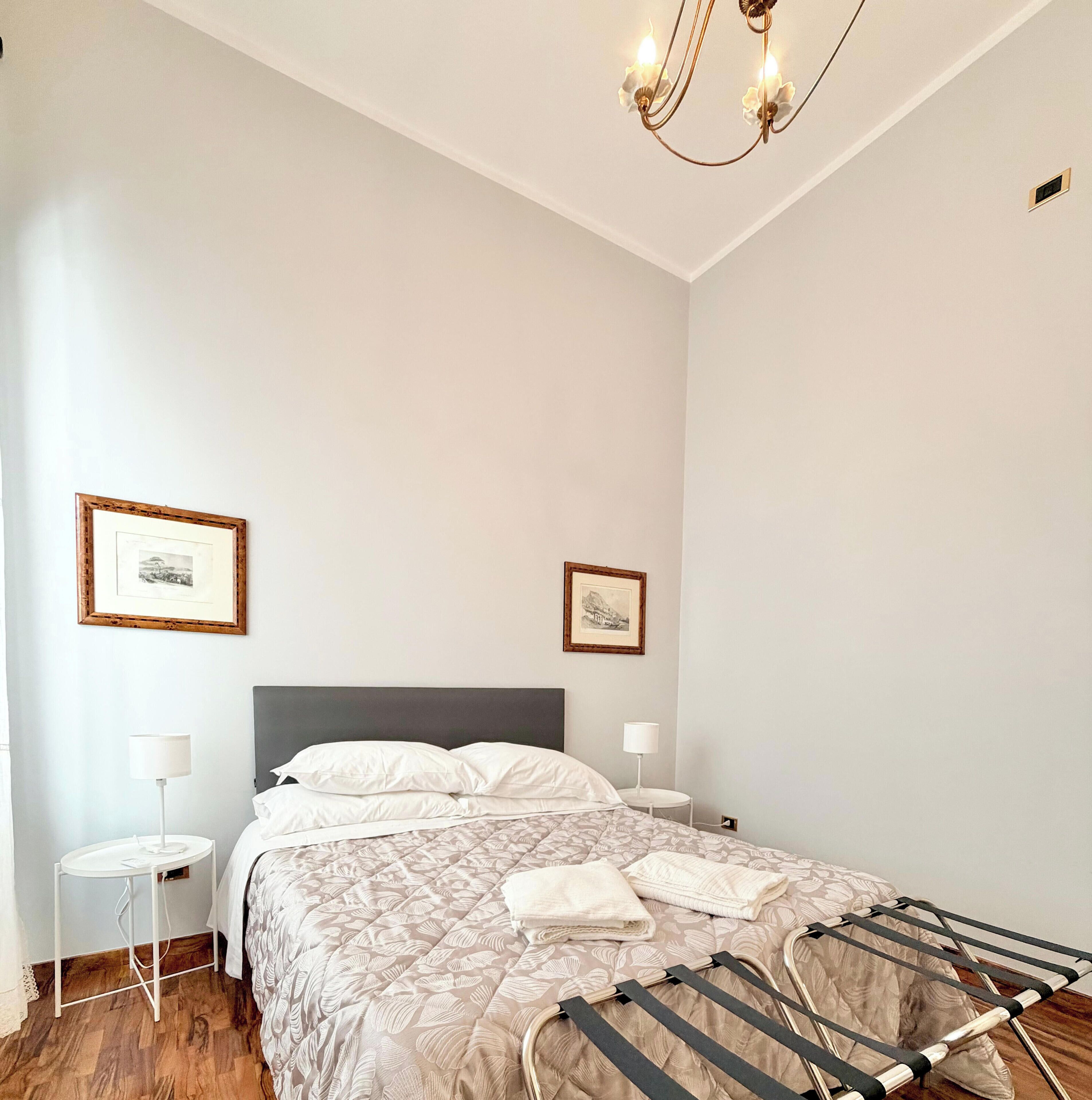 A bedroom with a bed, two bedside lamps, a chandelier, and two framed pictures on the wall.