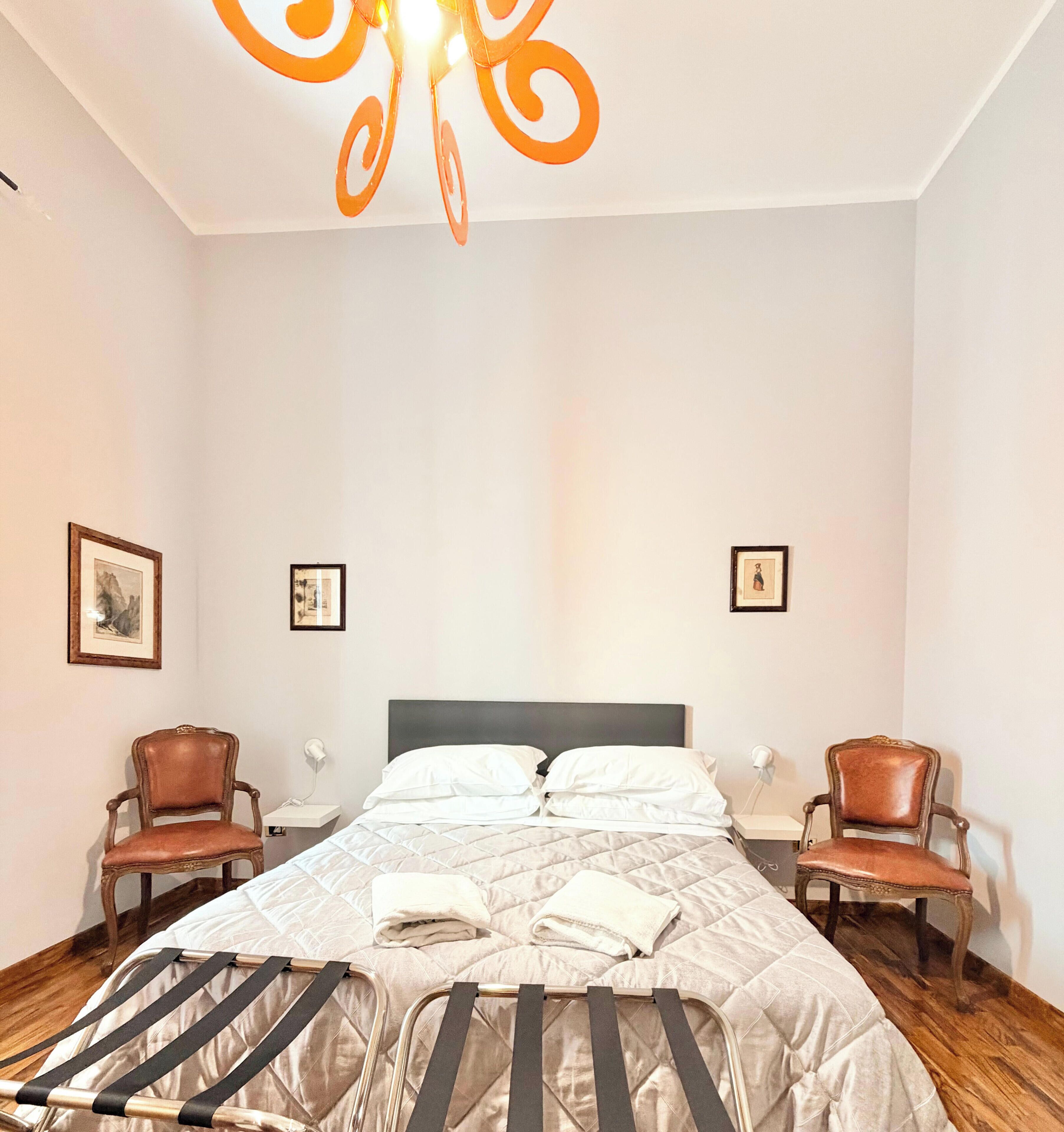 A hotel room with a bed, two chairs, a nightstand, and a ceiling light fixture.