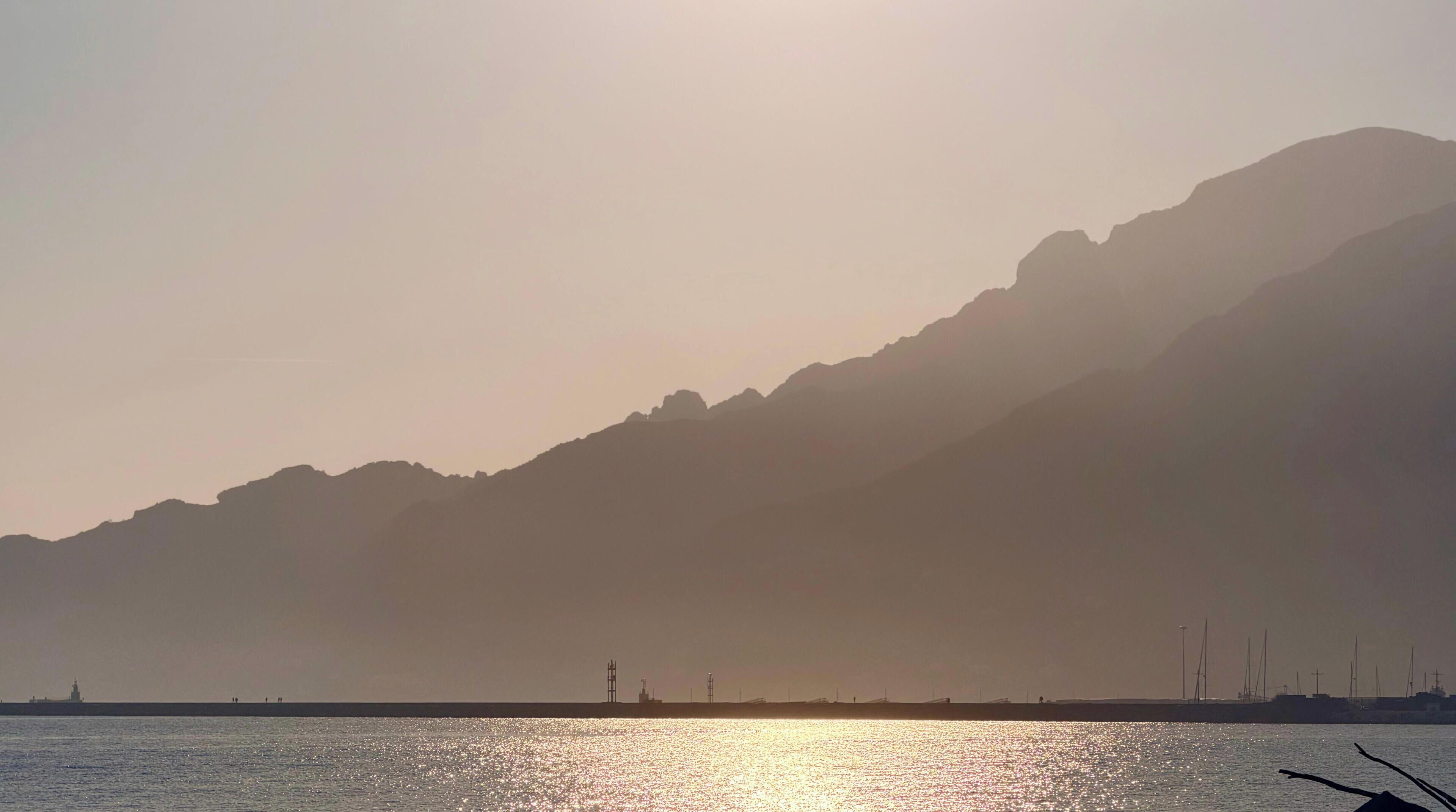 A coastal landscape with a mountain in the background and a calm sea reflecting the sunlight.