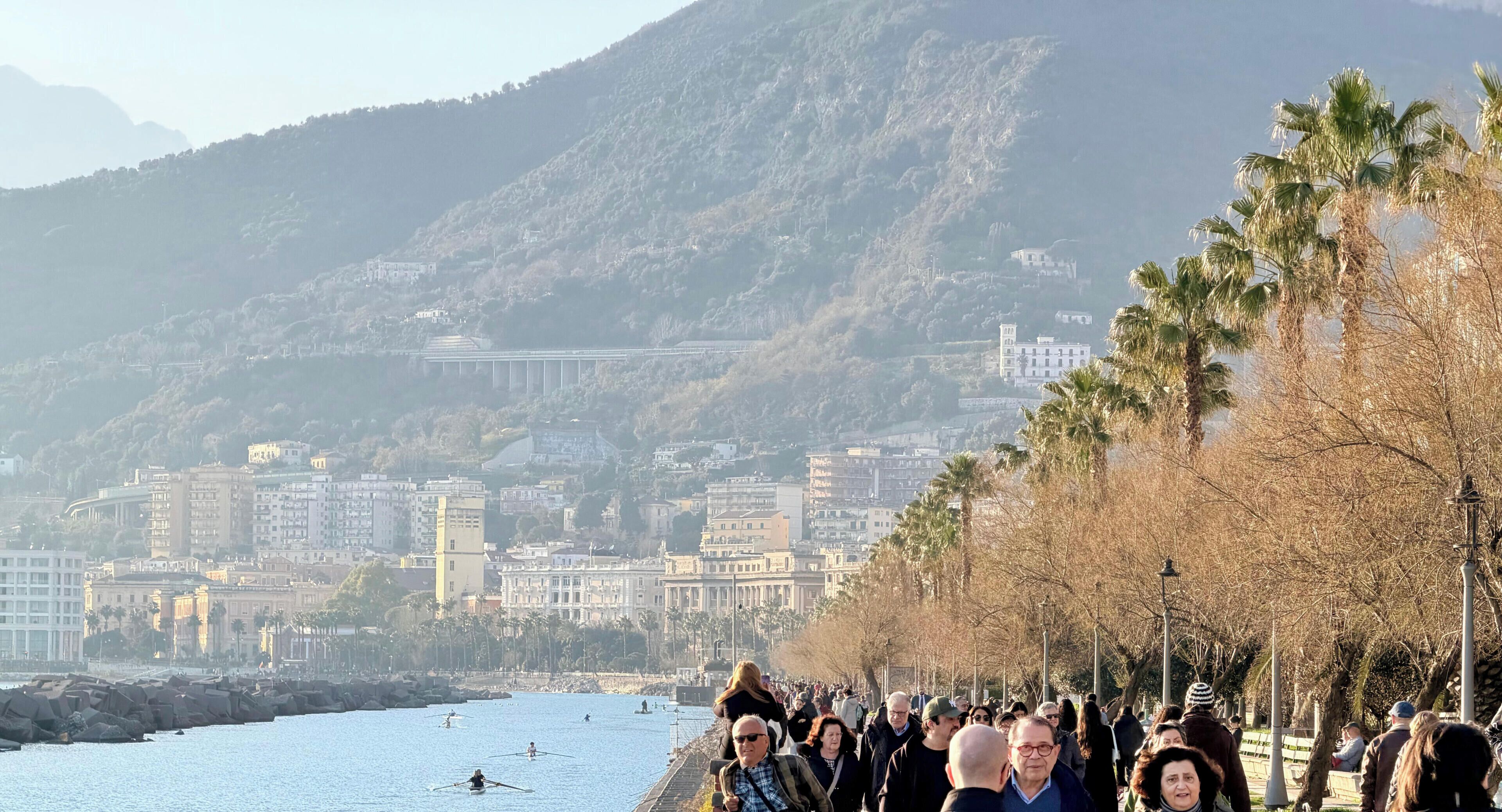 Un paseo marítimo con gente, una masa de agua y edificios al fondo.