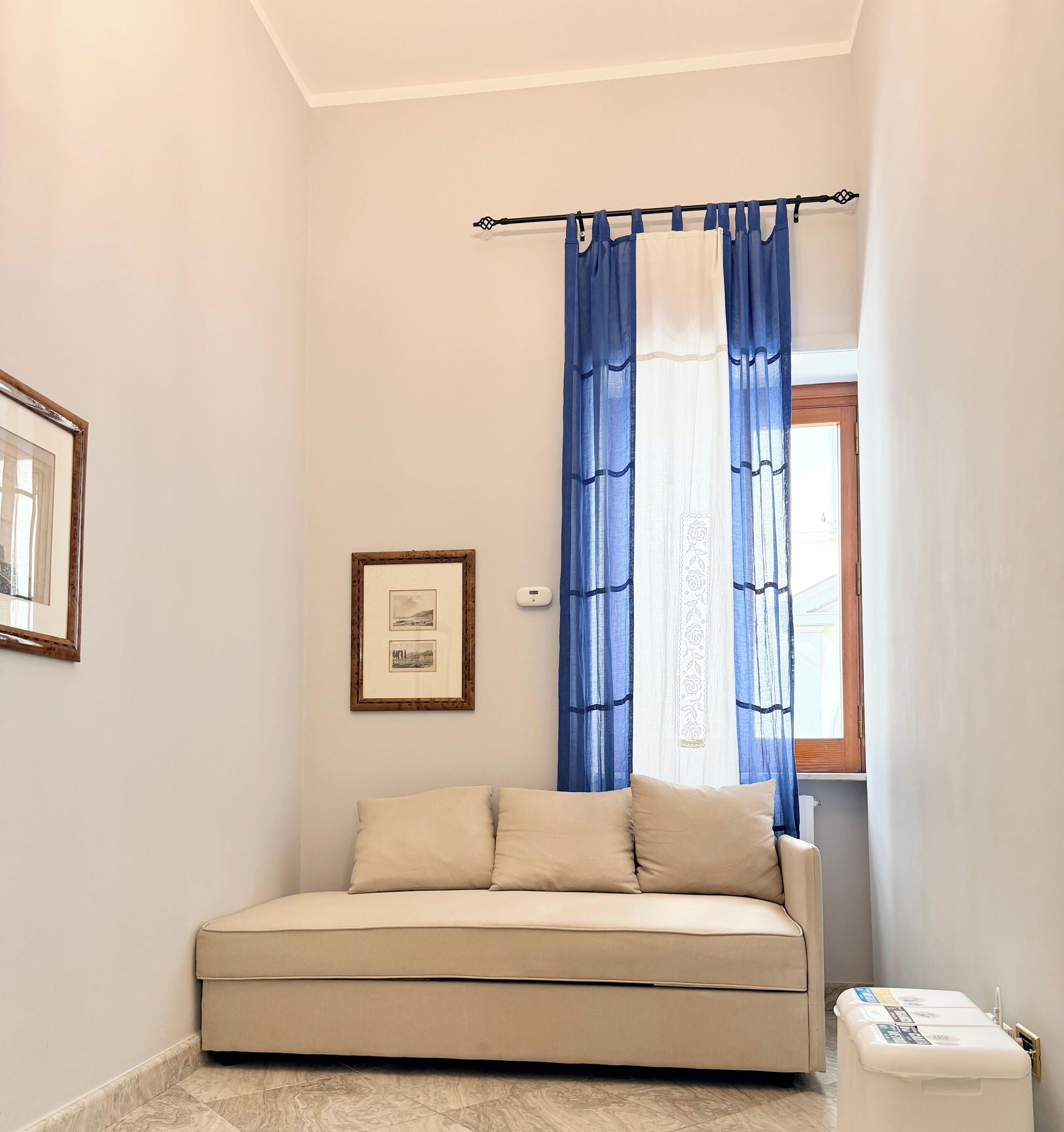 Une petite pièce avec un canapé beige, un tableau encadré au mur et une fenêtre ornée de rideaux bleus.