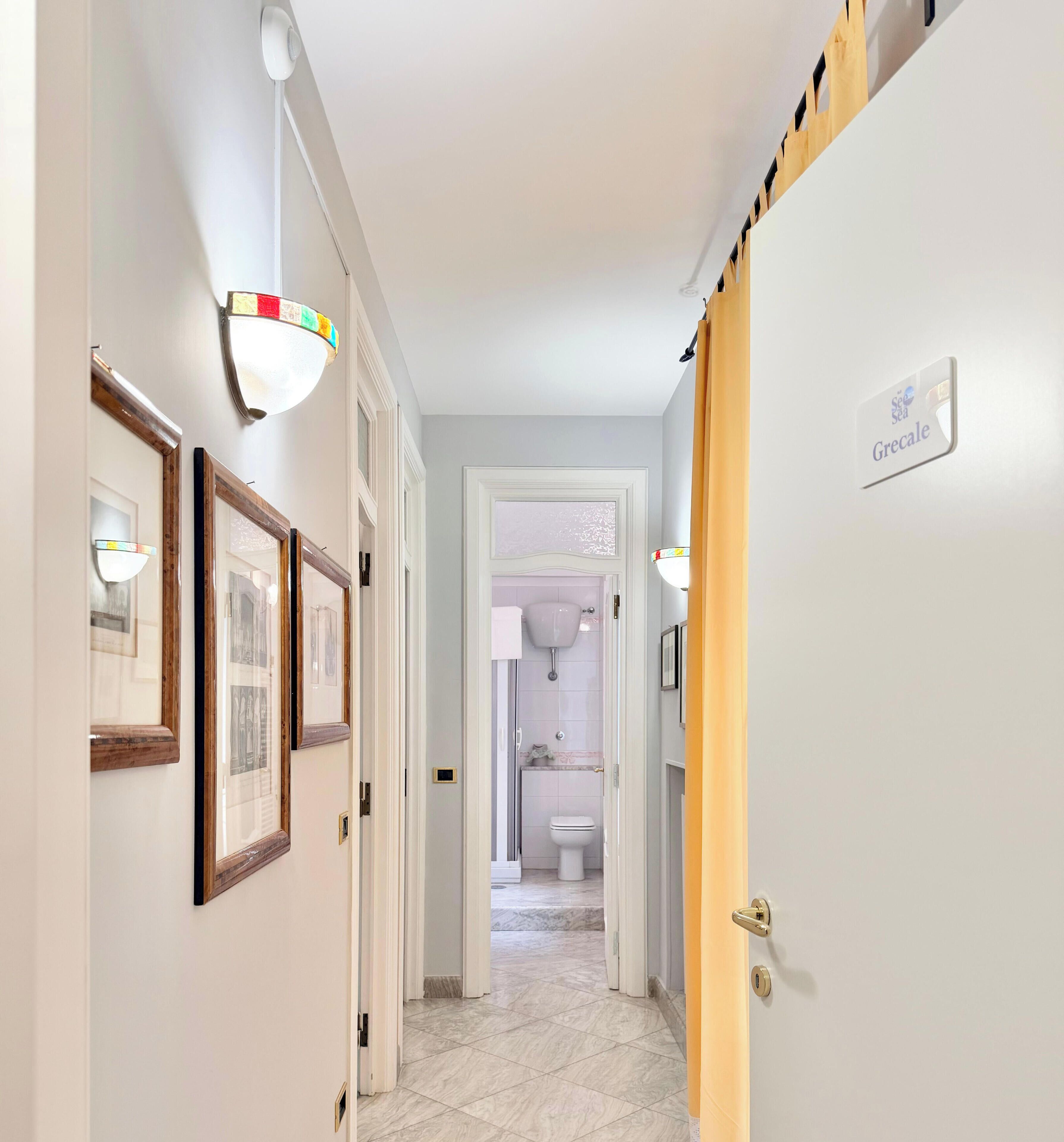 A hallway with framed pictures, a door labeled 'Sea Sea Grecale,' and a bathroom visible through an open door.