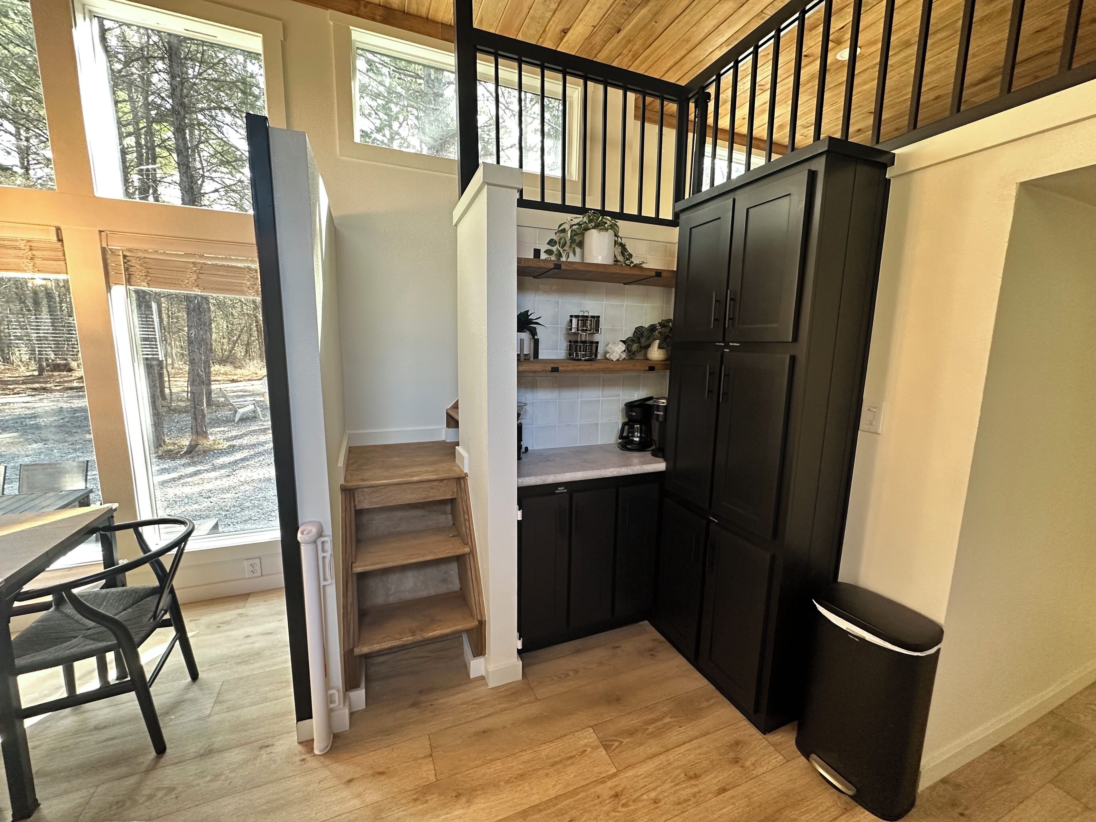 Une cuisine compacte avec des meubles de couleur sombre, un escalier en bois et une vue sur l’extérieur grâce à de grandes fenêtres.