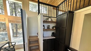 A compact kitchen with dark cabinets, a wooden staircase, and a view of the outdoors through large windows. - Cozy Cypress Cabin (Mount Vernon)