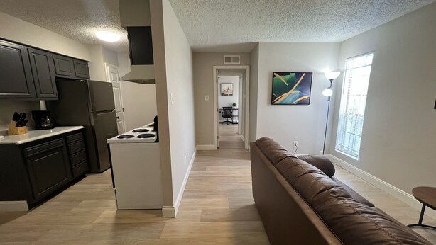 A modern kitchen with dark cabinets, a white stove, and a countertop with a coffee maker. A brown leather couch is in the foreground, and a painting is on the wall.