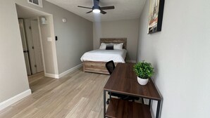 A bedroom with a bed, a ceiling fan, a wooden table with a plant, and a door leading to another room.