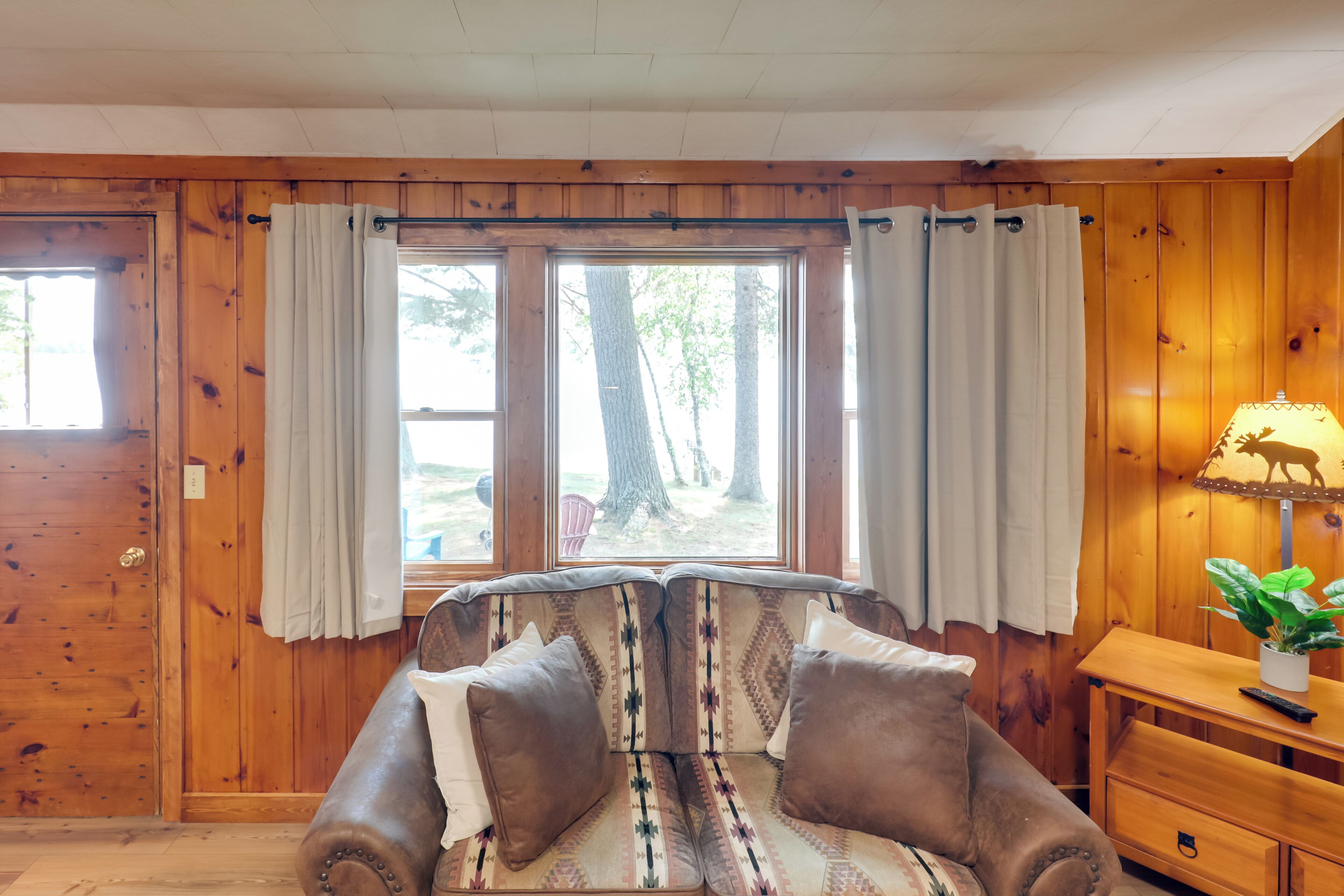A wooden-paneled room with a sofa, curtains, a lamp, and a view of trees outside.