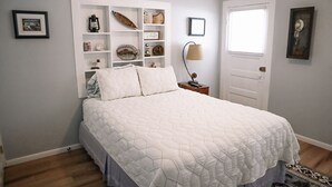 A bedroom with a bed, a bedside table with a lamp, a wall-mounted shelf with various items, and a door with a window.