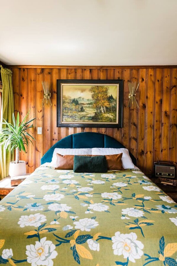 A bedroom with a wooden paneled wall, a framed landscape painting, a bed with a floral bedspread, a green plant, and a vintage radio.