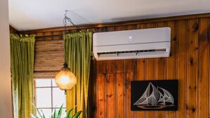 Ein gemütliches Zimmer mit einem mit grünem Stoff bezogenen Sofa, Holzboden, einer Topfpflanze, einer Pendelleuchte und einer wandmontierten Klimaanlage.
