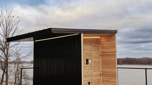Una pequeña cabaña de madera con techo negro, ubicada sobre una terraza de madera a orillas del agua.