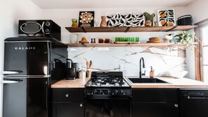A modern kitchen with a black refrigerator, stove, and dishwasher. There are wooden shelves above the counter holding various kitchen items and decorations.