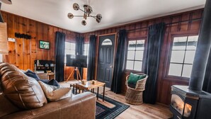A cozy living room with a leather sofa, a small table, a wicker chair, a fireplace, and wooden walls.