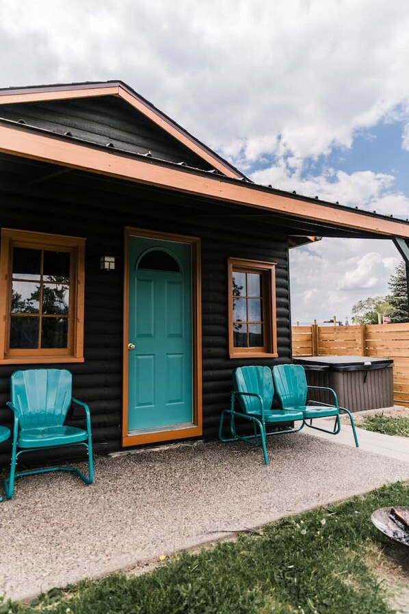 A log cabin with a teal door, two teal chairs, and a hot tub.