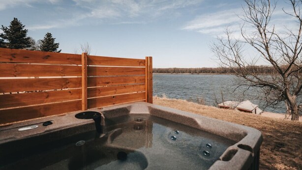 A hot tub with a wooden fence backdrop, a view of a lake, and a tree.