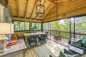 A wooden deck with a table and chairs, a hanging light fixture, and a view of trees through large windows. - Hike, Fish & Explore: Family Retreat in Highlands (Highlands)