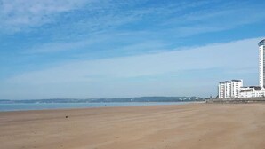 A wide sandy beach with a clear sky. In the background, there are high-rise buildings and a distant shoreline.