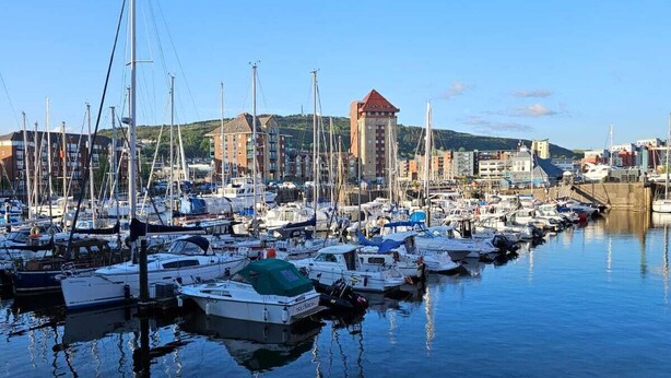 A marina with numerous boats docked, surrounded by buildings and hills in the background.