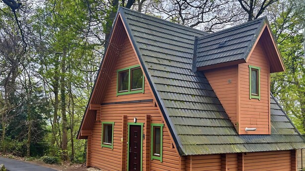 A wooden cabin with a steeply pitched roof, a front door, and two windows with green frames. - Acorn Lodge (Newton Abbot)