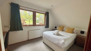 A bedroom with a large bed, a window with curtains, a radiator, and a bedside table with a lamp. - Acorn Lodge (Newton Abbot)