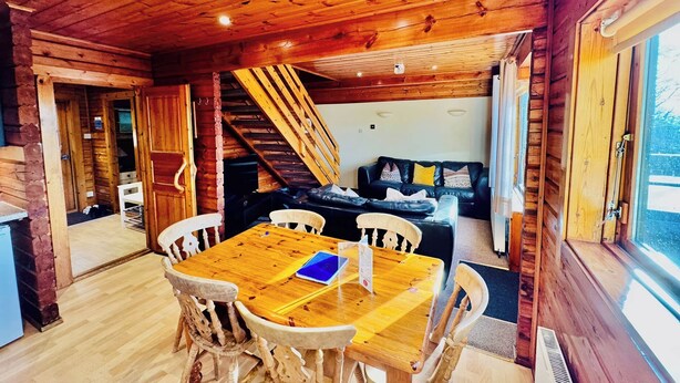 Interior de una cabaña de madera con una mesa de comedor, sillas, un sofá y una escalera.