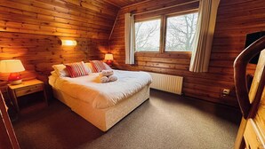 Un dormitorio acogedor tipo cabaña de madera con una cama, dos lámparas de noche, un ventanal con cortinas y un radiador.