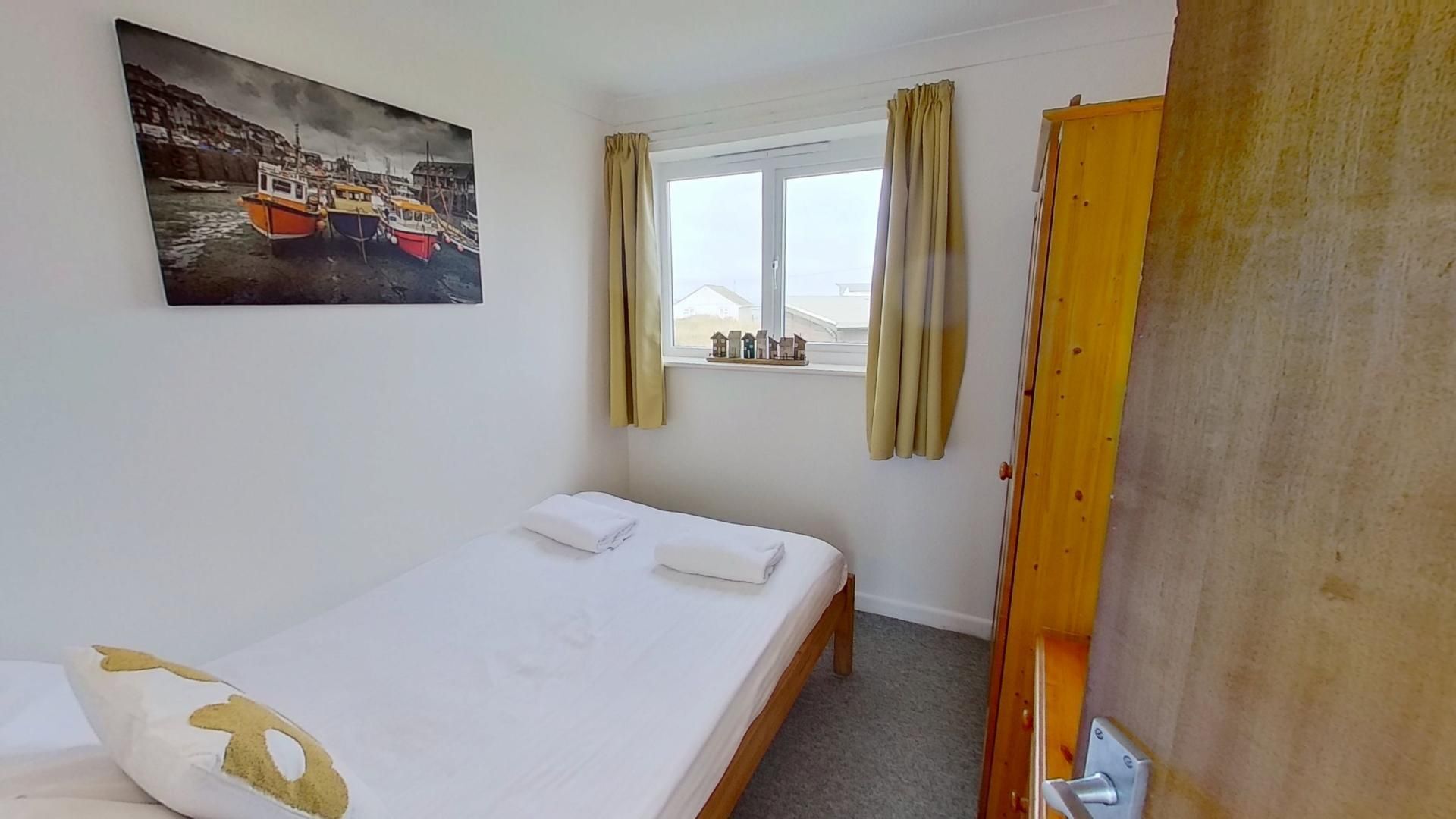 A small, simple room with a bed, a window with curtains, a painting of boats, and a door.