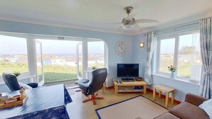 Ein geräumiges Wohnzimmer mit großen Fenstern, einem Deckenventilator, einem Flachbildfernseher und einem Essbereich mit Tisch und Stühlen.