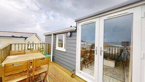 A wooden deck with a table and chairs, a sliding glass door, and a small window with a clock. - E41 Silver Mist, Riviere Towans (Hayle)