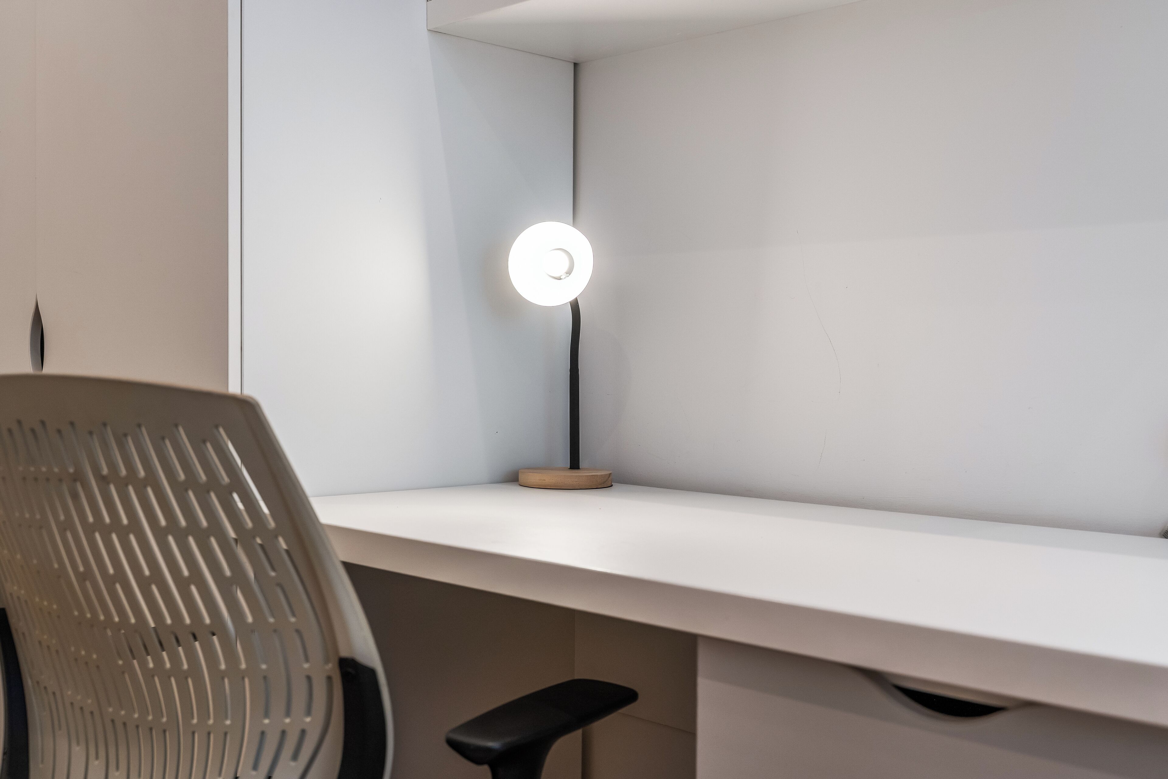 Un espacio de trabajo moderno con un escritorio blanco, una silla negra y una lámpara de pie con luz circular.
