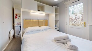 A neatly made bed with white linens and a folded towel, a bedside table, a window with a view of trees, and a door with a glass panel. - 58 Trevithick Court, Tolroy Manor (Hayle)